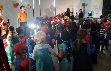 Várias dezenas de crianças em convívio em sala interior. Junto delas estão alguns adultos. Estão praticamente todos mascarados para o carnaval. Os rostos das crianças foram digitalmente desfocados.