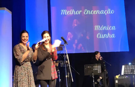 Fotografia de palco com várias pessoas a aplaudir. Ao fundo, em projeção, a expressão “Melhor Encenação: Mónica Cunha”.