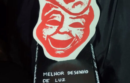 Plano aproximado de um troféu relativo ao “Melhor Desenho de Luz”. O troféu tem duas imagens estilizadas relativas a teatro e apresenta a expressão “Era uma vez... Teatro – Feitas de Ferro, Desenhadas a Carvão”.