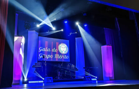 Fotografia de palco iluminado, mas sem pessoas. Em cima do palco estão já alguns instrumentos. Em projeção em tela, ao fundo, logótipo GDMA e expressão “Gala do Grupo Mérito”.