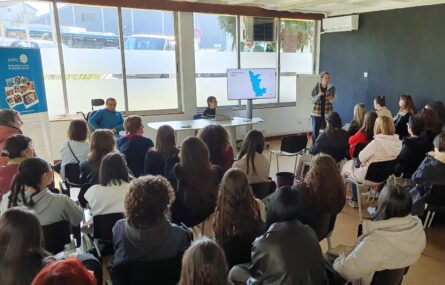 Mais de duas dezenas de pessoas sentadas, de costas, em sala interior. Ao fundo uma pessoa, de pé, faz uma apresentação oral com recurso a apresentação em televisor.