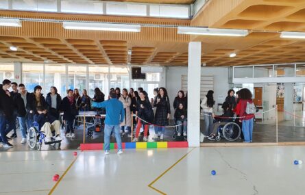 Duas dezenas de jovens (e adultos) em ginásio interior. No chão estão espalhadas algumas bolas de boccia (azuis, encarnadas e uma branca). Três pessoas usam cadeira de rodas.