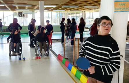 Fotografia de ginásio interior. Em primeiro plano uma pessoa segura uma raquete de boccia. Mais ao fundo outras pessoas conversam e preparam-se para a atividade.