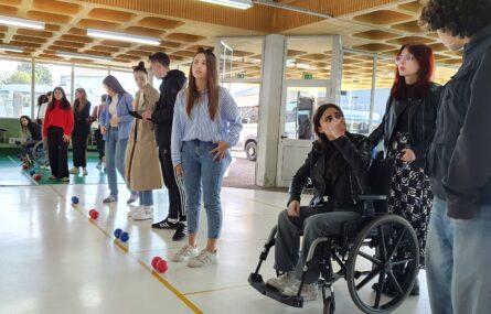 Cerca de uma dezena de pessoas em perspetiva diagonal. Preparam-se para uma partida de boccia estando pousadas, no chão, várias bolas azuis, encarnada e uma branca. Ao fundo são visíveis, pelas janelas, várias viaturas estacionadas.