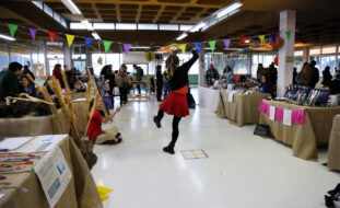 Fotografia de espaço interior com várias mesas com diversos produtos à venda. Várias pessoas transitam pelo espaço. Em destaque, ao centro, uma pessoa (de costas) aparenta dançar.