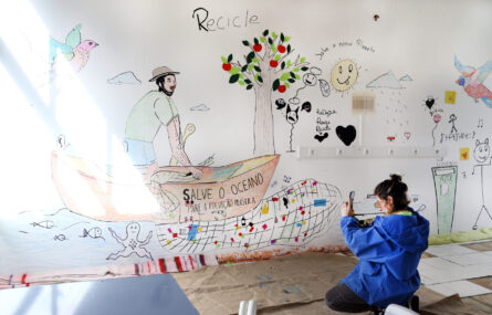 De joelhos uma pessoa usa o telemóvel para fotografar parede na qual foi pintado um mural (com diversas imagens, palavras e frases).
