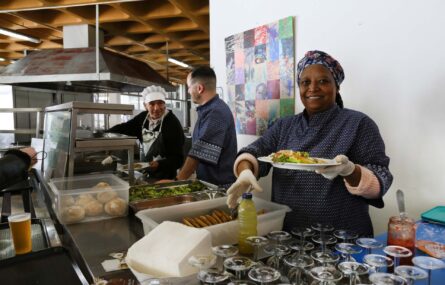 Três pessoas atrás de um balcão com vários produtos alimentares, copos e guardanapos. Uma das pessoas, sorridente, segura na mão esquerda um prato com comida.