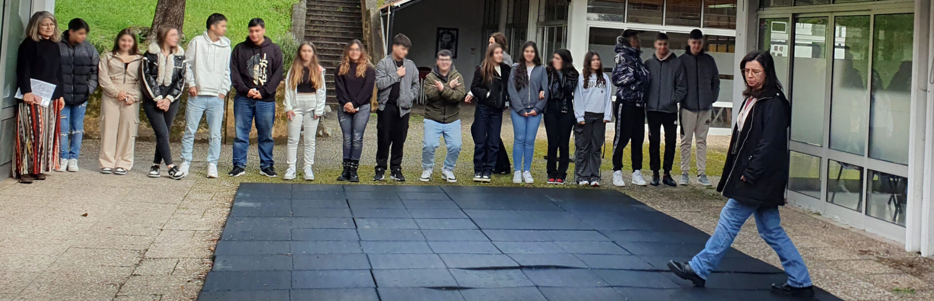 Grupo de 16 jovens lado a lado em espaço exterior. Junto deles estão três pessoas adultas. Ao fundo espaço ajardinado e alguns edifícios. Os rostos dos jovens foram digitalmente desfocados.