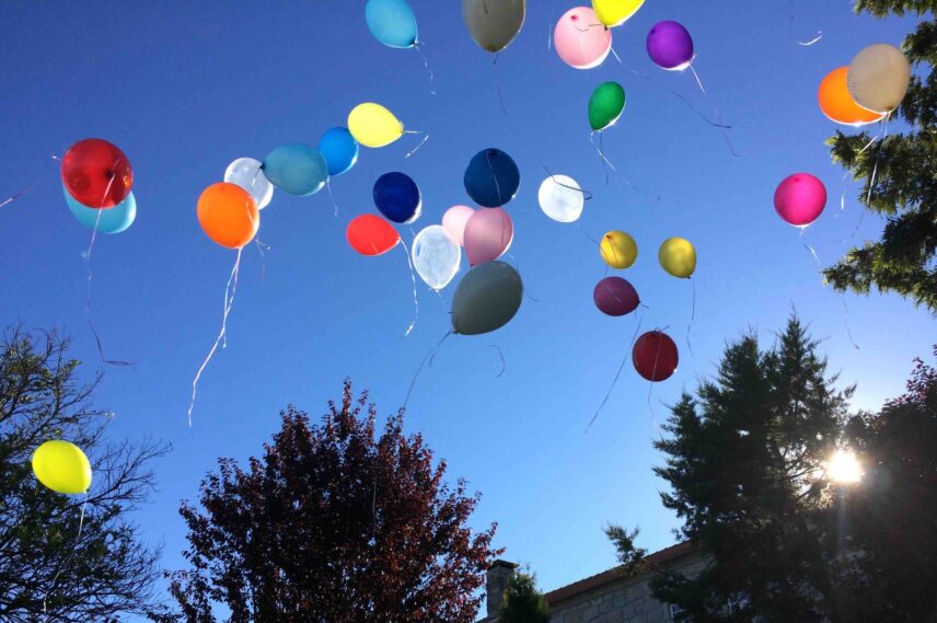 Largada de diversos balões coloridos. São lançados para o céu num dia de sol. Algumas árvores aparecem no enquadramento.