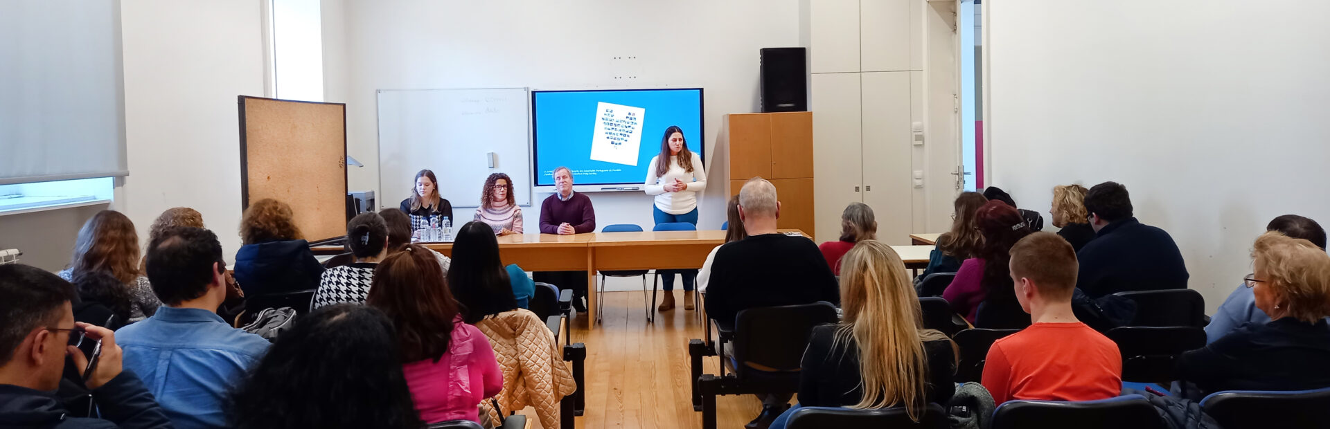Fotografia de sala interior. De costas, sentadas em cadeiras, estão cerca de duas dezenas de pessoas. Ao fundo, junto de duas mesas compridas, estão quatro pessoas. Uma das pessoas, de pé, faz apresentação oral com recurso a projeção em ecrã.