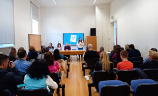Fotografia de sala interior. De costas, sentadas em cadeiras, estão cerca de duas dezenas de pessoas. Ao fundo, junto de duas mesas compridas, estão quatro pessoas. Uma das pessoas, de pé, faz apresentação oral com recurso a projeção em ecrã.
