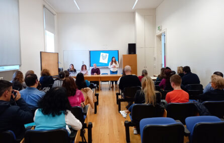 Fotografia de sala interior. De costas, sentadas em cadeiras, estão cerca de duas dezenas de pessoas. Ao fundo, junto de duas mesas compridas, estão quatro pessoas. Uma das pessoas, de pé, faz apresentação oral com recurso a projeção em ecrã.