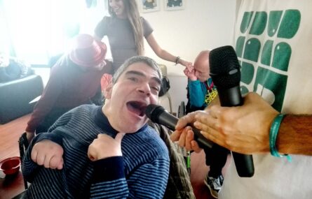 Uma pessoa está a cantar – usando um microfone que está na mão de outra pessoa, a seu lado. Ao fundo mais três pessoas em atividade.