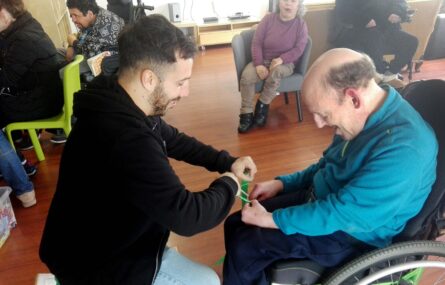 Duas pessoas, sorridentes, tentam fazer algo com uma fita verde que seguram nas mãos. Ao fundo da imagem outras pessoas em atividade.