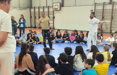 Fotografia de interior de pavilhão com várias dezenas de jovens e adultos. No centro do círculo formado pelos jovens estão duas pessoas realizar uma explicação oral. Os rostos visíveis dos jovens estudantes foram digitalmente desfocados.