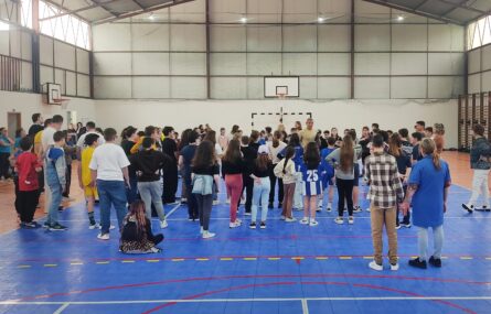 Fotografia de interior de pavilhão com várias dezenas de jovens e adultos. Os rostos visíveis dos jovens estudantes foram digitalmente desfocados.