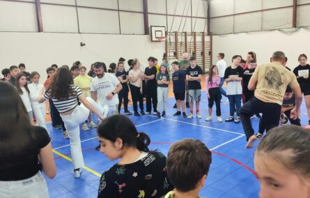 Fotografia de interior de pavilhão com várias dezenas de jovens e adultos. No centro do círculo formado pelos jovens estão quatro pessoas a praticar Capoeira. Os rostos dos jovens estudantes foram todos digitalmente desfocados.