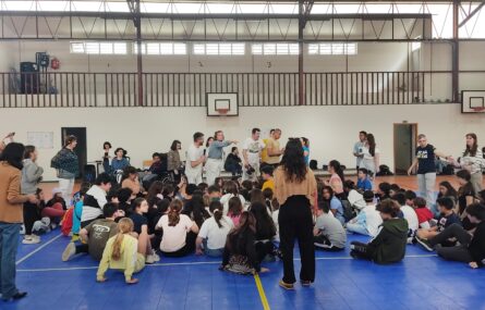 Fotografia de interior de pavilhão com várias dezenas de jovens e adultos. Os rostos visíveis dos jovens estudantes foram digitalmente desfocados.