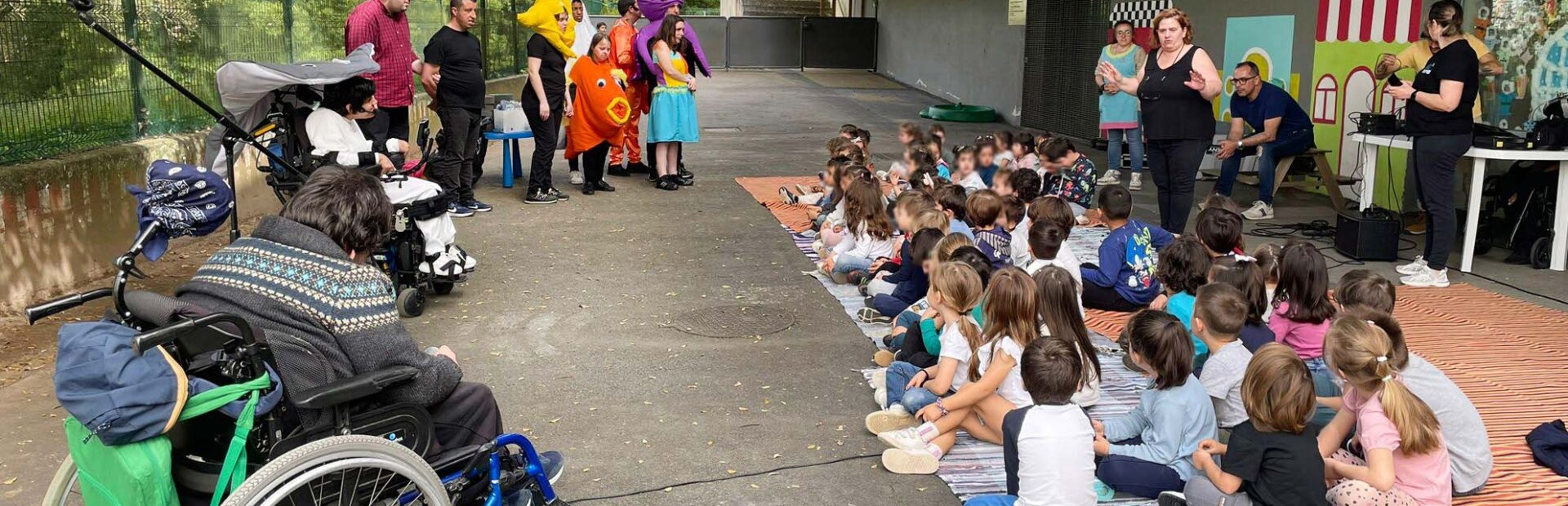 Fotografia de espaço exterior. Várias dezenas de crianças, sentadas no chão, assistem a uma apresentação de elementos de um grupo de teatro. Ao fundo da imagem, do lado esquerdo, várias árvores. Os rostos das crianças foram digitalmente desfocados.