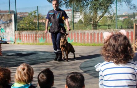 De costas várias crianças assistem a uma atividade feita por uma pessoa adulta acompanhada por um cão (ou cadela...) que está preso por uma trela.