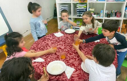 Sete crianças junto de uma mesa. Algumas delas estão a brindar com os copos cheios (do que aparenta ser sumo de laranja). Os rostos das crianças foram digitalmente desfocados.
