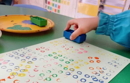 Plano aproximado da mão de uma criança que usa uma peça de lego, invertida, para carimbar uma folha de papel com círculos de várias cores.