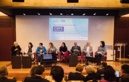 Plano geral de oito pessoas sentadas no palco (participantes do painel “Prescrição Social em Portugal – experiências de implementação”, em projeção de ecrã). De costas algumas pessoas nas primeiras filas da assistência.