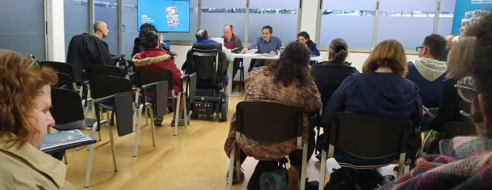Várias pessoas sentadas em sala interior. Ao fundo uma mesa com três pessoas lado a lado. Em primeiro plano uma pessoa, de costas, consulta vários documentos.