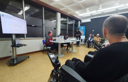 Três pessoas sentadas junto de uma mesa repleta de documentos. Mais ao fundo, de pé, uma pessoa faz uma apresentação oral. Em primeiro plano, de costas, pessoa sentada em cadeira de rodas.