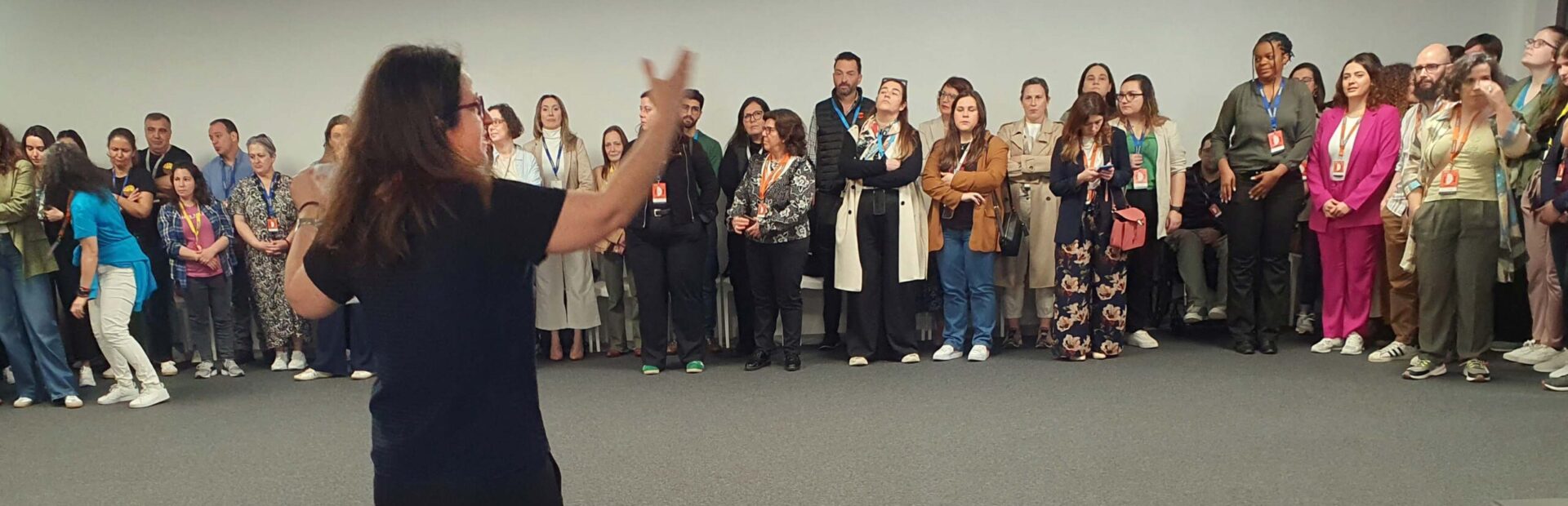 De costas uma pessoa fala e gesticula com muitas outras pessoas (alinhadas ao fundo da imagem). Em primeiro plano, pousados numa mesa, um computador portátil, um microfone e um projetor de vídeo.
