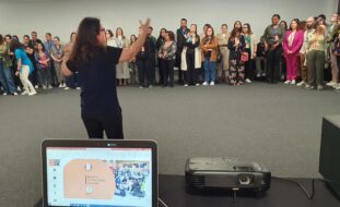 De costas uma pessoa fala e gesticula com muitas outras pessoas (alinhadas ao fundo da imagem). Em primeiro plano, pousados numa mesa, um computador portátil, um microfone e um projetor de vídeo.