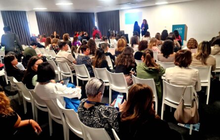 Várias dezenas de pessoas sentadas, de costas, em cadeiras brancas. Ao fundo um pequeno palco com duas pessoas em conversa.