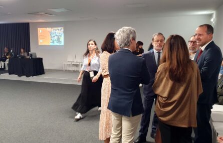 Do lado direito da imagem está um grupo de seis pessoas em conversa. Ao centro há uma outra pessoa a falar. Ao fundo da imagem um pequeno palco com uma projeção vídeo na parede.