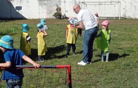 Perto de uma dezena de crianças, com um adulto, estão a jogar futebol em espaço relvado. Os rostos visíveis de algumas das crianças foram digitalmente desfocados.