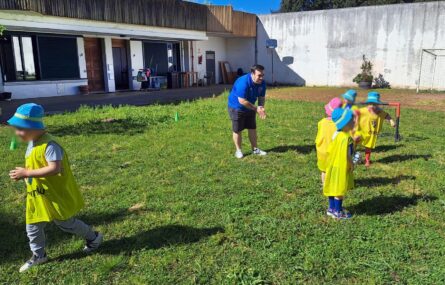 Cinco crianças e um adulto, estão a jogar futebol em espaço relvado exterior. Os rostos visíveis de algumas das crianças foram digitalmente desfocados.
