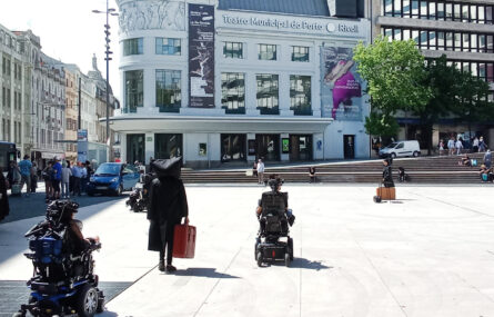 Vários atores em ação de rua num espaço público exterior. Ao fundo alguns edifícios, viaturas e pessoas a transitar e a assistir à apresentação.
