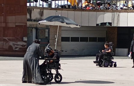 Vários atores em ação de rua num espaço público exterior. Ao fundo alguns espaços comerciais, viaturas e pessoas a transitar e a assistir à apresentação.