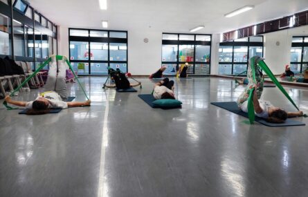 Cerca de uma dezena de pessoas em atividade física em ginásio. Todas estão deitadas em tapetes dispostos no chão e elevam as pernas.