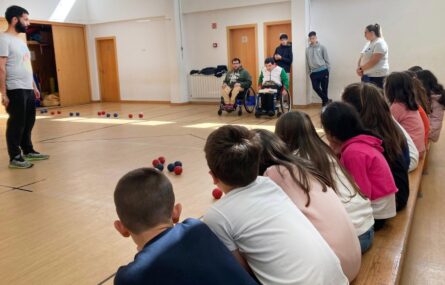 Fotografia de pequeno pavilhão interior. Cerca de uma dezena de crianças assiste a uma demonstração de boccia. Dois praticantes em cadeira de rodas estão junto de várias bolas (azuis e vermelhas) espalhadas pelo chão. Os rostos das crianças foram digitalmente desfocados.