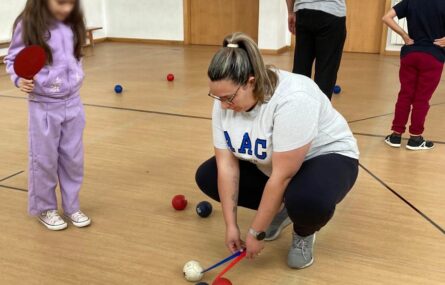Uma criança verifica a medição que uma pessoa adulta faz da distância entre várias bolas de boccia (espalhadas no chão do pavilhão). A criança tem o rosto digitalmente desfocado. Ao fundo da imagem, de costas, outros dois praticantes.