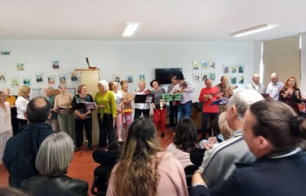 Em primeiro plano algumas pessoas de costas. Ao fundo da imagem, e já de frente, grupo de 18 pessoas que aparentam estar a cantar. Algumas dessas pessoas estão a utilizar instrumentos musicais. A parede mais ao fundo da imagem está repleta de quadros com rostos de pessoas.