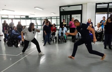 Duas pessoas praticam capoeira. Atrás e ao lado cerca de duas dezenas de pessoas estão a assistir e a bater palmas.
