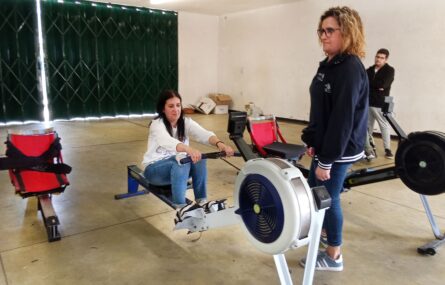 Uma pessoa está a usar um equipamento desportivo para prática de remo. De pé está uma pessoa a apoiar e, ao fundo da imagem, uma outra pessoa assiste.