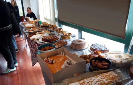Mesa repleta de doces, bolos e aperitivos. Ao fundo da imagem algumas pessoas em conversa.