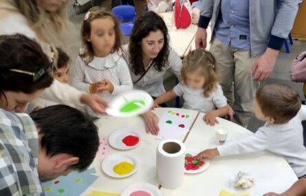 Cerca de uma dezena de pessoas, adultos e crianças, junto de uma mesa. Estão a fazer pinturas em cartolinas. Os rostos das crianças foram digitalmente desfocados.
