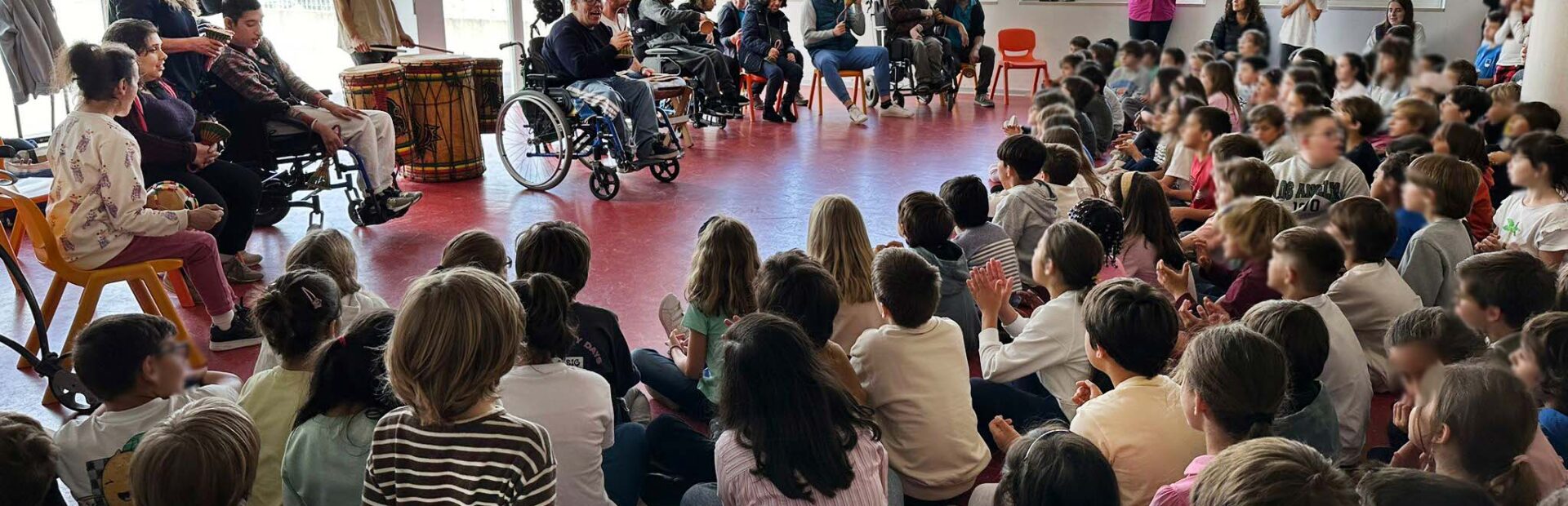 Fotografia de espaço interior de escola. Várias dezenas de crianças sentadas no chão a assistir a uma apresentação feita por um grupo de jovens e adultos. Os rostos visíveis das crianças foram digitalmente desfocados.