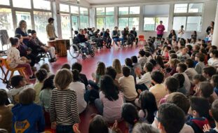 Fotografia de espaço interior de escola. Várias dezenas de crianças sentadas no chão a assistir a uma apresentação feita por um grupo de jovens e adultos. Os rostos visíveis das crianças foram digitalmente desfocados.