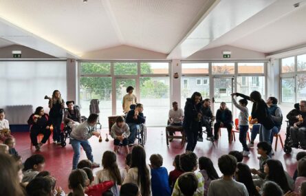Fotografia de espaço interior de escola. Várias dezenas de crianças sentadas no chão a assistir a uma apresentação feita por um grupo de jovens e adultos. Algumas crianças, jovens e adultos aparentam estar a dançar. Outros usam vários instrumentos musicais. Os rostos visíveis das crianças foram digitalmente desfocados.