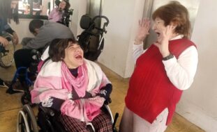 Em primeiro plano duas pessoas claramente sorridentes. Mais ao fundo outras pessoas.