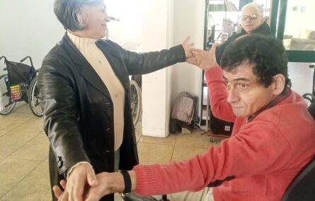 Duas pessoas aparentam estar a dançar (encontrando-se de mãos dadas). Mais ao fundo outra pessoa assiste.